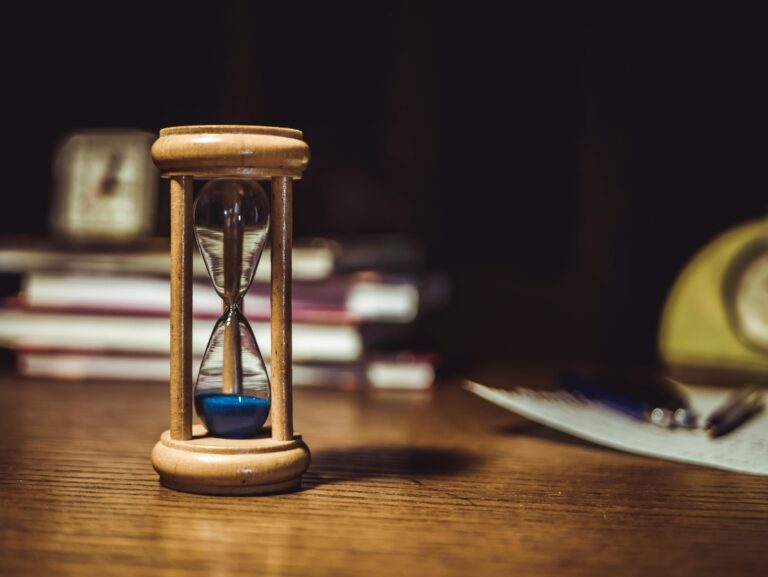 Wooden hourglass with blue sand on a desk beside notebooks and a pen, symbolizing reflection and the time it takes to explain.